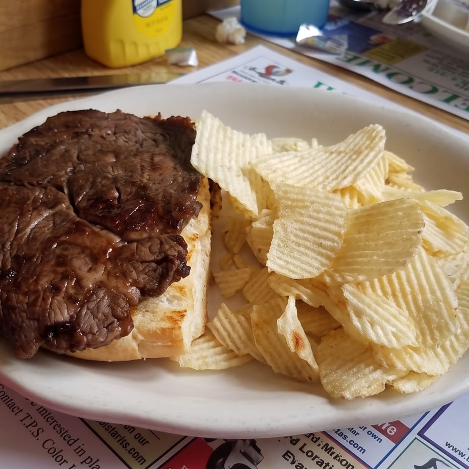 A steak sandwich with a side of ruffled potato chips on a white plate at a casual restaurant.