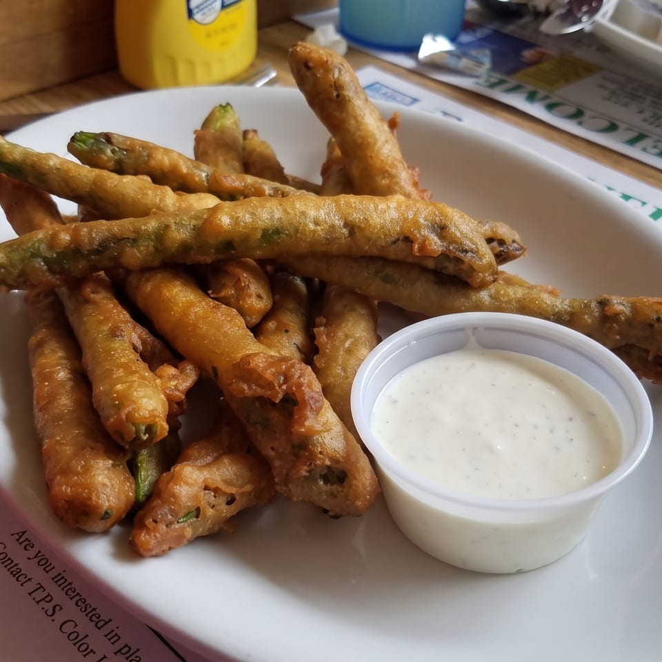 A white plate holds a pile of golden-brown fried green beans with a side of ranch dressing.