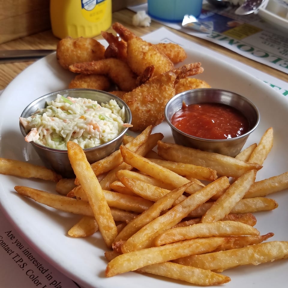 A plate of fried shrimp, french fries, coleslaw, and ketchup on a table with a blue cup.