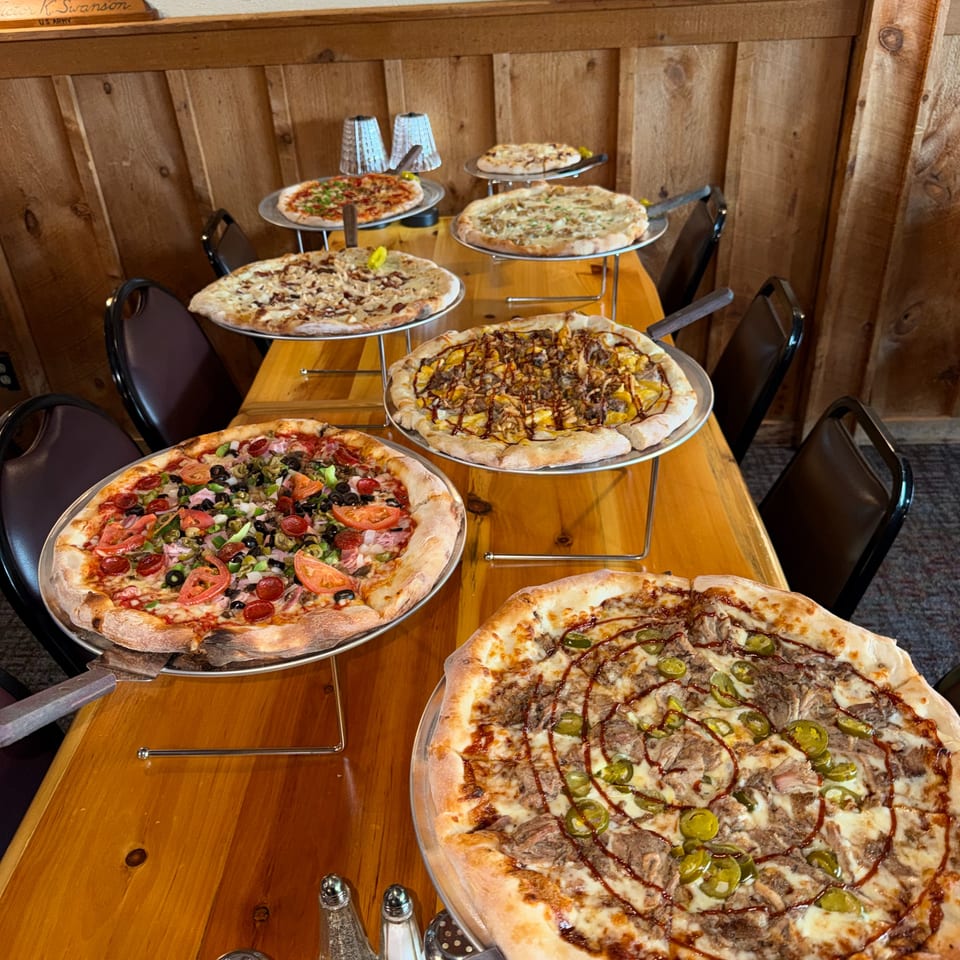 Assorted gourmet pizzas displayed on elevated stands along a wooden restaurant table beneath a folded American flag.